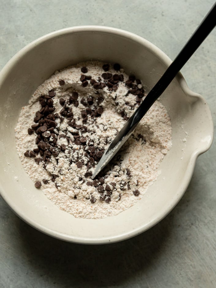 Overhead view of a cream ceramic mixing bowl containing flour and chocolate chips being stirred with a black spatula, showing the dry ingredients before wet ingredients are added.