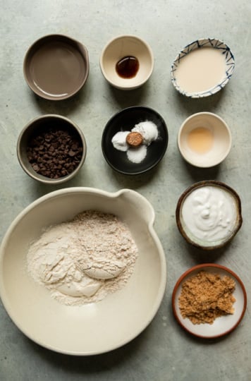Overhead flat lay of all the muffin ingredients measured out in individual small bowls on a grey surface, including all-purpose and whole wheat flours, chocolate chips, plant-based milk, vanilla extract, coconut milk or cream, brown sugar, leavening agents, salt, nutmeg, oil, and vegan yogurt.