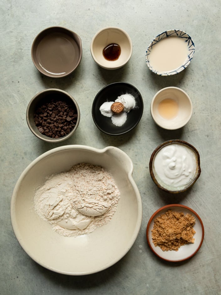 Overhead flat lay of all the muffin ingredients measured out in individual small bowls on a grey surface, including all-purpose and whole wheat flours, chocolate chips, plant-based milk, vanilla extract, coconut milk or cream, brown sugar, leavening agents, salt, nutmeg, oil, and vegan yogurt.