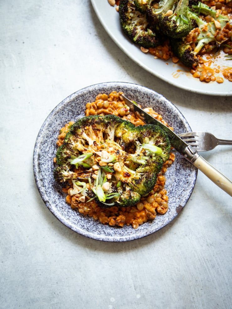 A grilled broccoli steak served over spicy peanut lentils in a speckled blue and white ceramic bowl, garnished with sliced green onions and chopped peanuts. A vintage-handled fork and knife rest on the rim. A second plated serving is visible in the upper right corner.