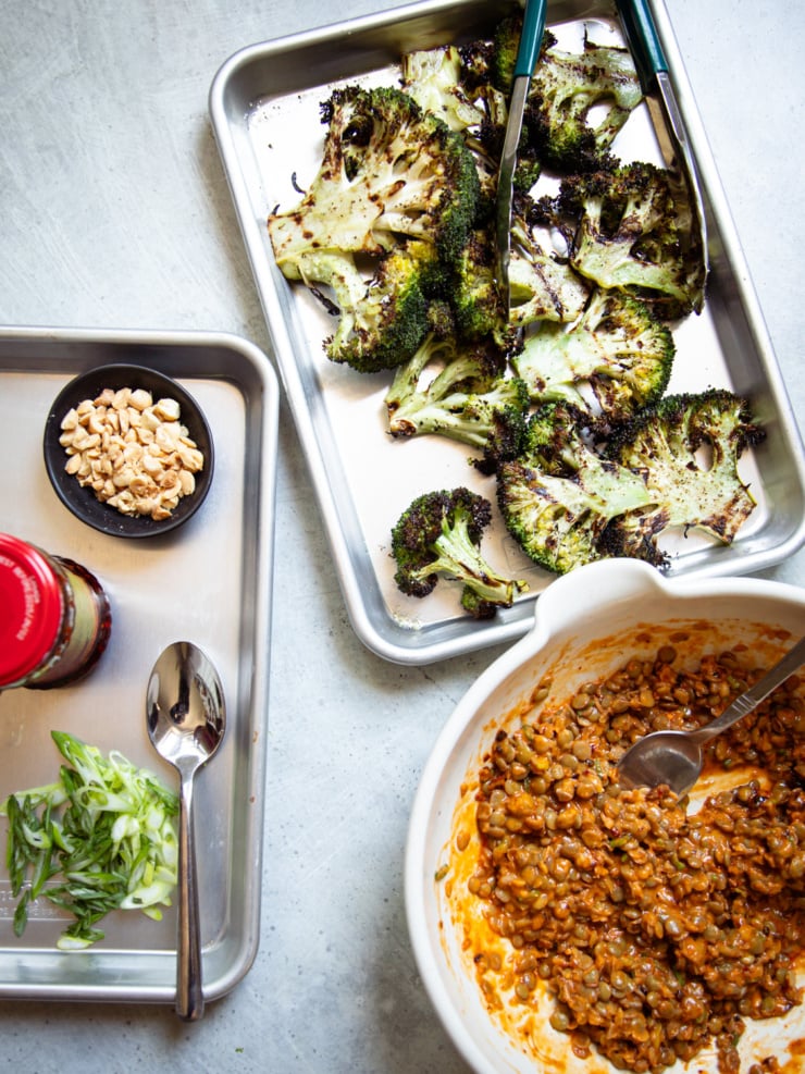 A flat lay of recipe components mid-assembly: a baking sheet of grilled broccoli steaks with teal-handled tongs, a white bowl of saucy lentils, and a small side tray holding chopped peanuts, sliced green onions, a spoon, and a jar of chili crisp.