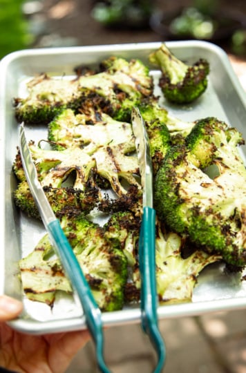 Close-up of a hand holding a baking sheet of freshly grilled broccoli steaks with deep char marks, teal tongs resting on top, with a lush green garden visible in the background.