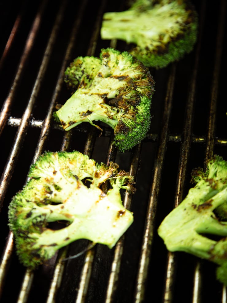 A dark, moody close-up of broccoli steaks on a hot grill grate, showing deep char marks and caramelization on the florets and stems.