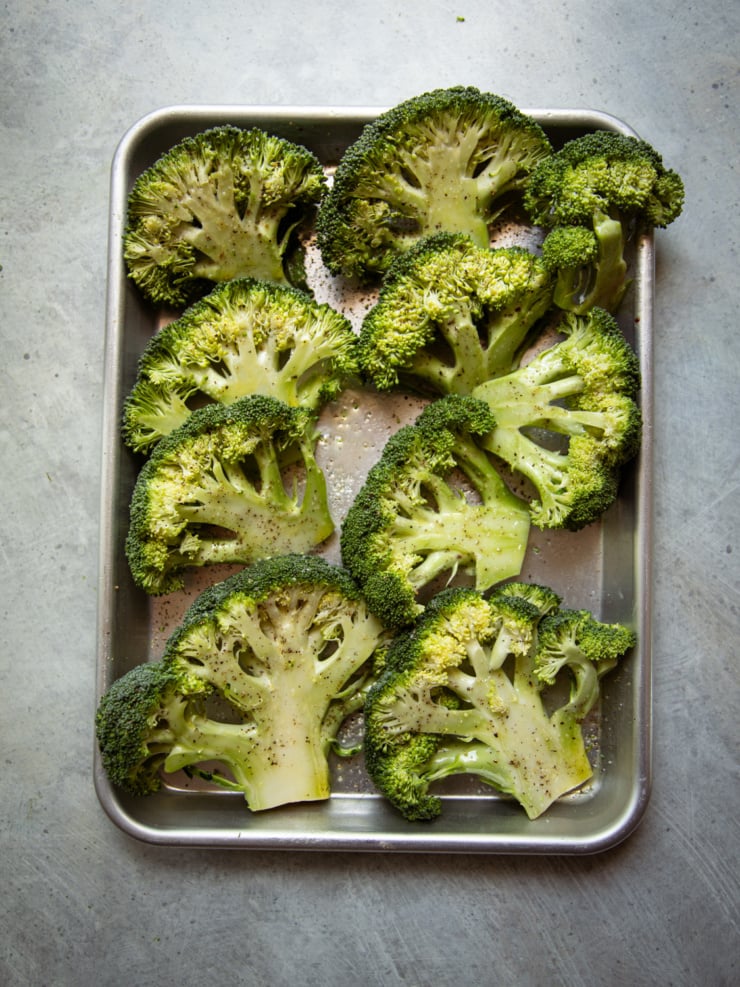 Overhead shot of raw broccoli steaks — thick cross-section slices — laid flat on a silver baking sheet, oiled and seasoned with salt and pepper, ready for the grill.