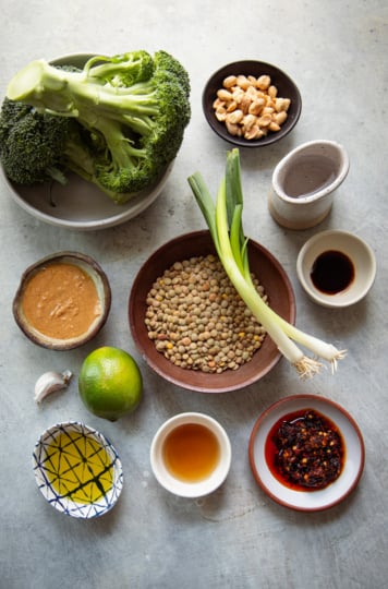 A flat lay ingredients shot featuring whole broccoli, dry green lentils, green onions, peanut butter, lime, garlic, chili crisp, tamari, maple syrup, avocado oil, and roasted peanuts, arranged in various small bowls and vessels on a light grey surface.