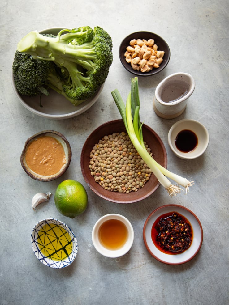 A flat lay ingredients shot featuring whole broccoli, dry green lentils, green onions, peanut butter, lime, garlic, chili crisp, tamari, maple syrup, avocado oil, and roasted peanuts, arranged in various small bowls and vessels on a light grey surface.