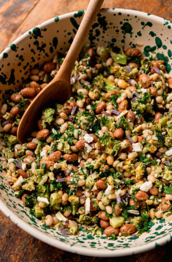 A close-up overhead shot of a finished lemony bean salad in a cream and green speckled bowl, with a wooden spoon resting inside. Navy beans and romano beans are visible throughout, mixed with chopped roasted broccoli, fresh parsley, crumbled vegan feta, and small purple chive blossoms as garnish.