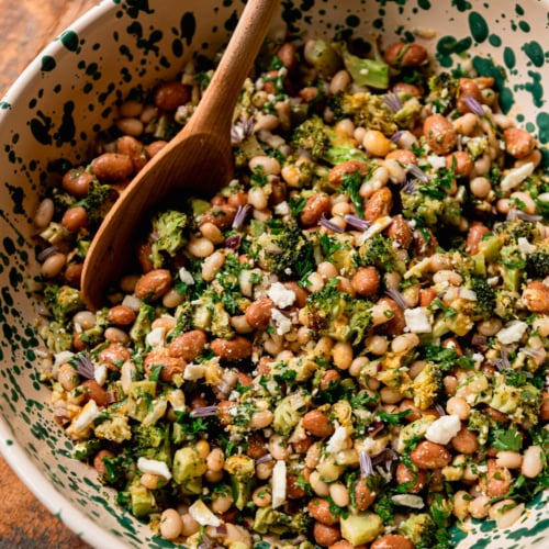 A close-up overhead shot of a finished lemony bean salad in a cream and green speckled bowl, with a wooden spoon resting inside. Navy beans and romano beans are visible throughout, mixed with chopped roasted broccoli, fresh parsley, crumbled vegan feta, and small purple chive blossoms as garnish.