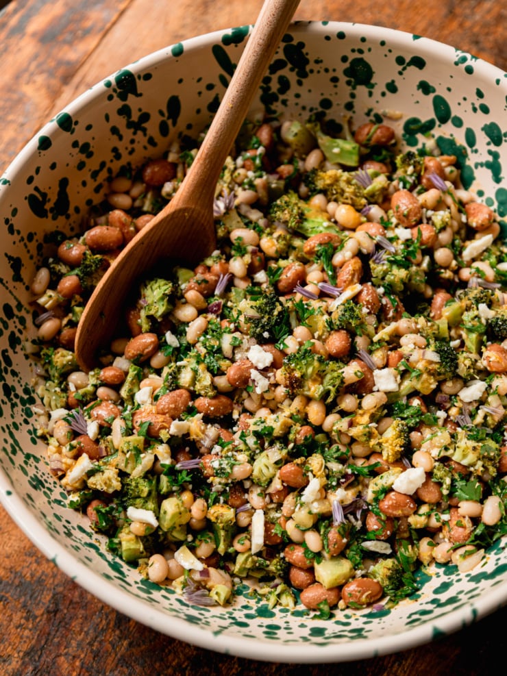 A close-up overhead shot of a finished lemony bean salad in a cream and green speckled bowl, with a wooden spoon resting inside. Navy beans and romano beans are visible throughout, mixed with chopped roasted broccoli, fresh parsley, crumbled vegan feta, and small purple chive blossoms as garnish.