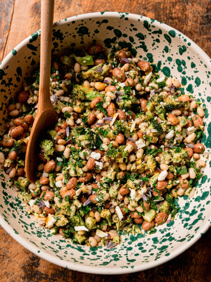 A slightly wider overhead view of a lemony bean salad in a green-speckled bowl with wooden spoon. The mix of navy and romano beans, roasted broccoli pieces, parsley, and vegan feta crumbles is clearly visible, garnished with scattered purple chive blossoms.