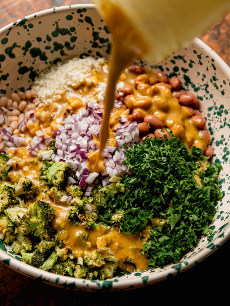 An action shot of golden-orange nutritional yeast dressing being poured from a small vessel into the speckled bowl, which holds the salad components arranged in sections: roasted broccoli, diced red onion, navy beans, romano beans, and a large mound of chopped parsley.