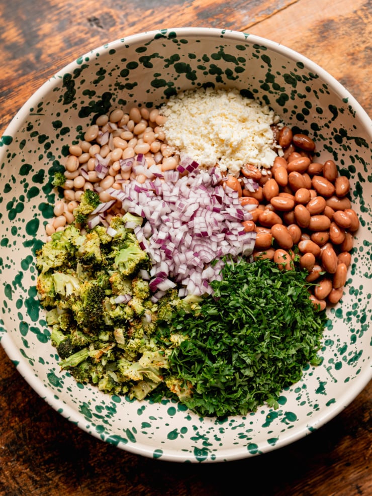An overhead flat-lay of the salad components arranged in the green-speckled bowl before mixing: roasted broccoli florets, finely diced red onion, crumbled vegan feta, navy beans, romano beans, and chopped fresh parsley.