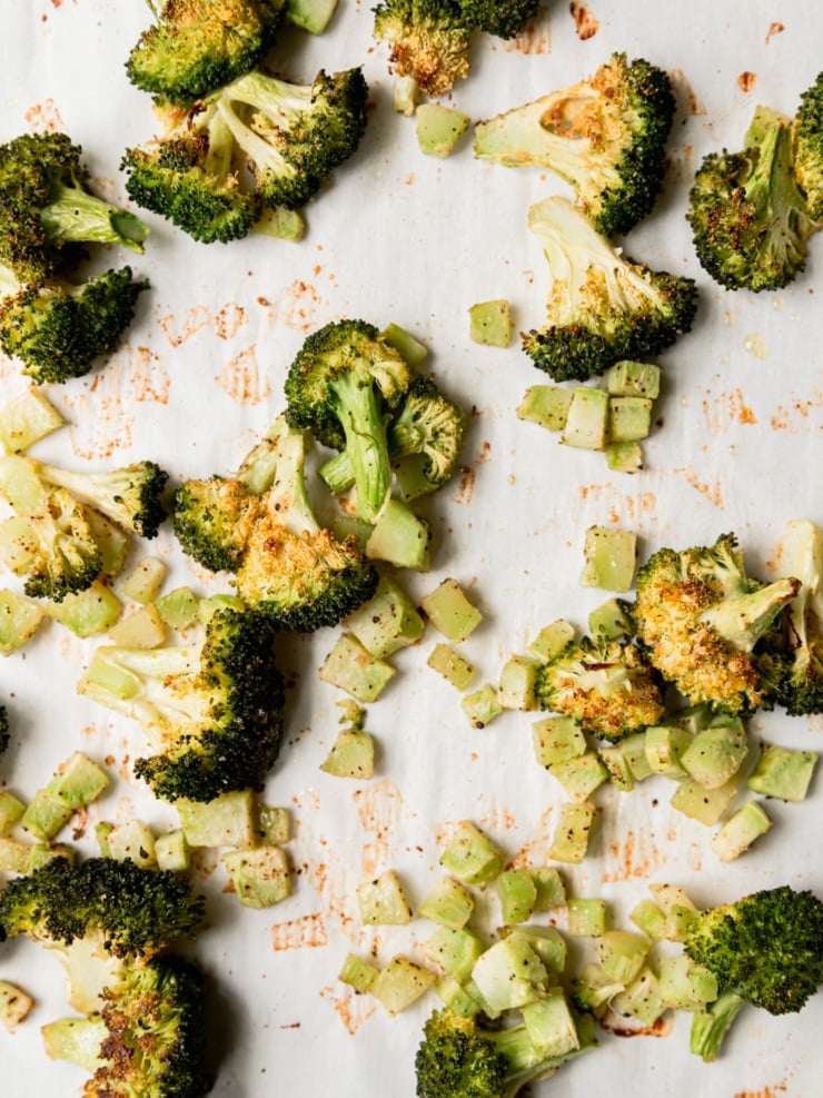 An overhead close-up of roasted broccoli florets and diced broccoli stems spread across a parchment-lined baking sheet, golden and caramelized at the edges with seasoning visible.
