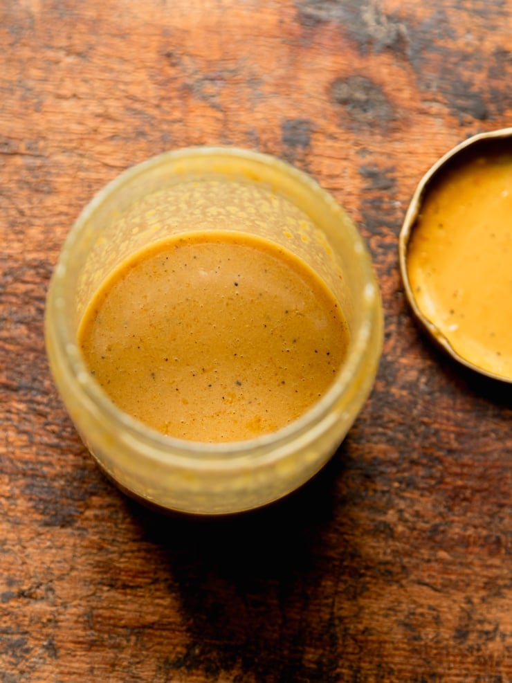 An overhead shot of the finished nutritional yeast dressing in an open glass jar, smooth and golden-orange in color, with the jar lid resting beside it on a dark wood surface.