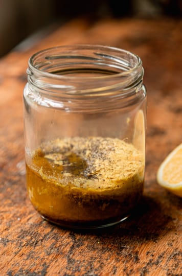 A close-up of a small glass jar on a wood surface holding the unmixed dressing ingredients — olive oil, lemon juice, nutritional yeast, and black pepper — with a halved lemon visible to the right.