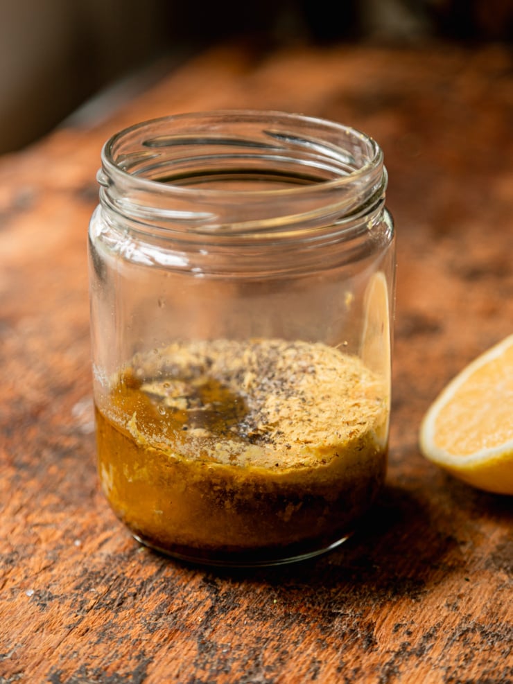 A close-up of a small glass jar on a wood surface holding the unmixed dressing ingredients — olive oil, lemon juice, nutritional yeast, and black pepper — with a halved lemon visible to the right.