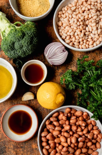 A flat-lay of all the raw recipe ingredients on a dark wood surface: a head of broccoli, a bowl of nutritional yeast, a bowl of navy beans, a halved red onion, olive oil, a small bowl of vegan Worcestershire sauce, a garlic clove, a lemon, fresh flat-leaf parsley, a second small bowl of maple syrup, and a bowl of romano beans.