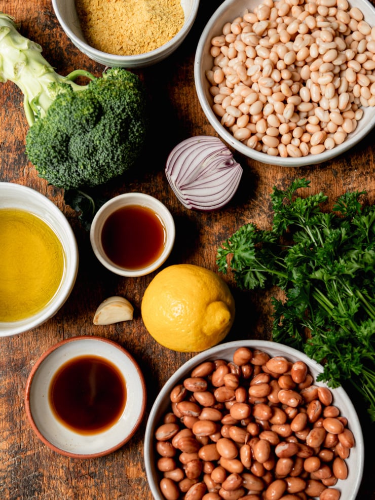 A flat-lay of all the raw recipe ingredients on a dark wood surface: a head of broccoli, a bowl of nutritional yeast, a bowl of navy beans, a halved red onion, olive oil, a small bowl of vegan Worcestershire sauce, a garlic clove, a lemon, fresh flat-leaf parsley, a second small bowl of maple syrup, and a bowl of romano beans.