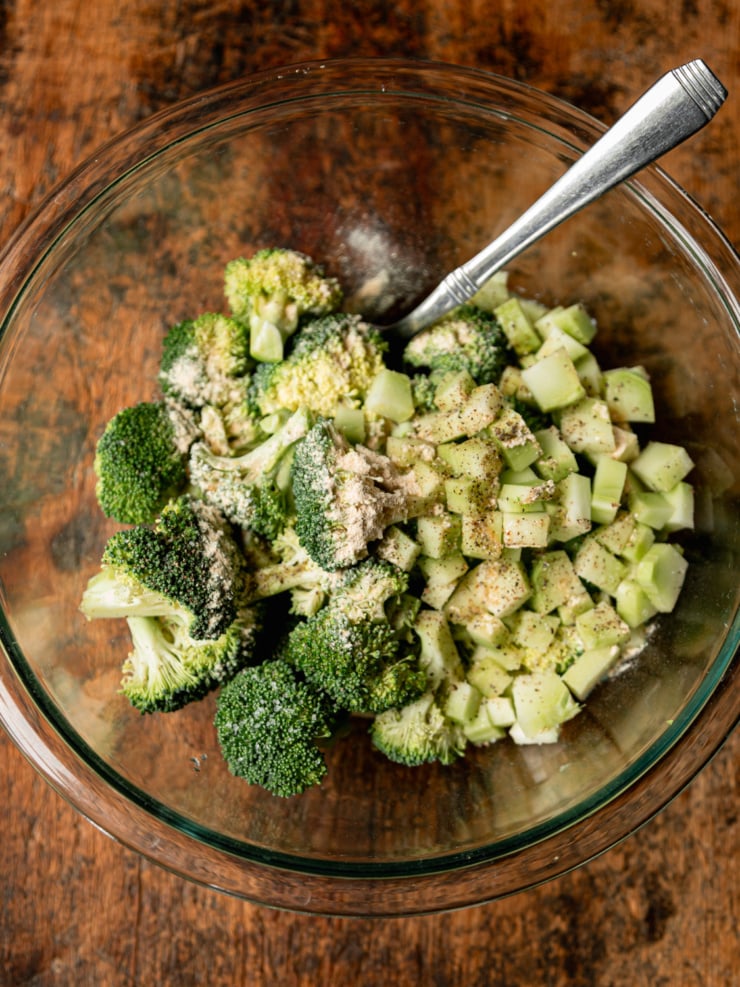 An overhead shot of raw broccoli florets and diced broccoli stems in a clear glass bowl, tossed with garlic powder, olive oil, salt, and black pepper before roasting, with a silver spoon resting inside.
