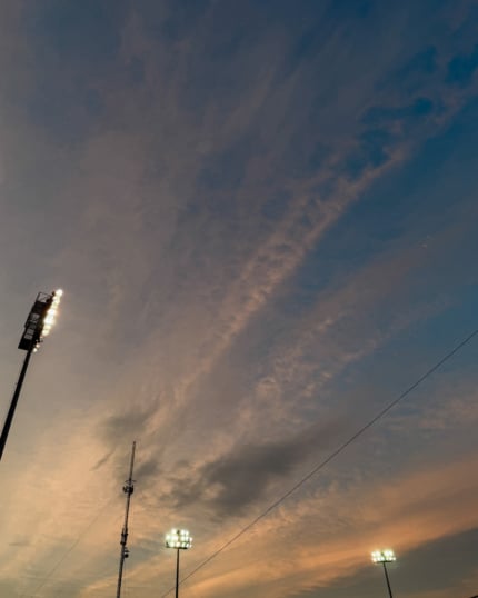 A dramatic dusk sky in shades of blue, grey, and warm amber, with wispy cirrus clouds streaking diagonally across the frame. Three illuminated stadium light towers rise from the bottom of the image against the colourful sky.