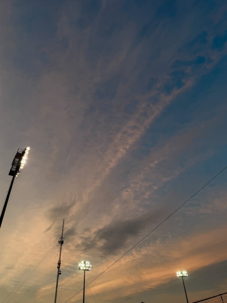 A dramatic dusk sky in shades of blue, grey, and warm amber, with wispy cirrus clouds streaking diagonally across the frame. Three illuminated stadium light towers rise from the bottom of the image against the colourful sky.