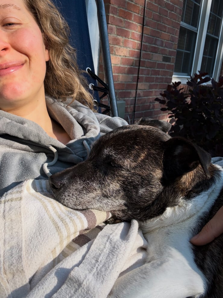 A smiling woman in a grey hoodie lounges in an outdoor chair, holding a dark brindle senior dog nestled against her chest. A red brick building and dark-leafed shrub are visible in the background.