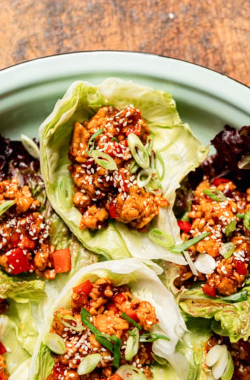 Close-up overhead shot of spicy sesame tempeh lettuce wraps nestled in iceberg and red leaf lettuce cups on a mint-green enamel plate, garnished with sliced green onions and white sesame seeds. Diced red bell pepper and glossy dark sauce are visible throughout the crumbled tempeh filling.