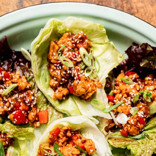 Close-up overhead shot of spicy sesame tempeh lettuce wraps nestled in iceberg and red leaf lettuce cups on a mint-green enamel plate, garnished with sliced green onions and white sesame seeds. Diced red bell pepper and glossy dark sauce are visible throughout the crumbled tempeh filling.