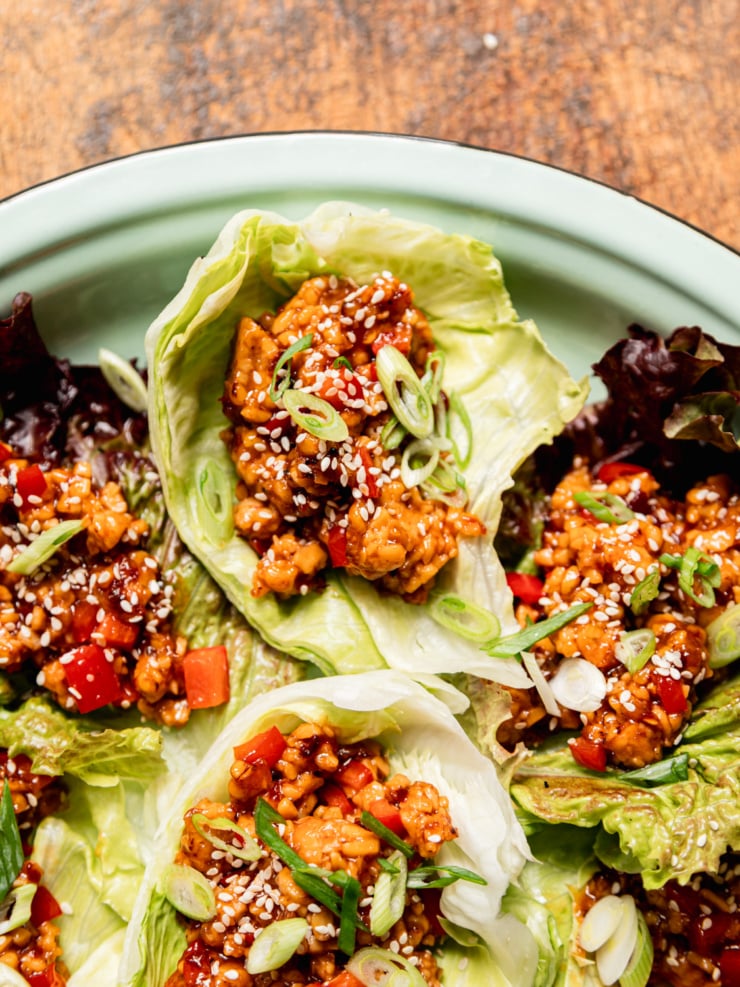 Close-up overhead shot of spicy sesame tempeh lettuce wraps nestled in iceberg and red leaf lettuce cups on a mint-green enamel plate, garnished with sliced green onions and white sesame seeds. Diced red bell pepper and glossy dark sauce are visible throughout the crumbled tempeh filling.