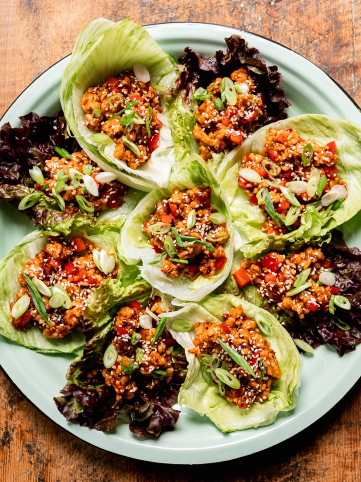 Overhead shot of a full mint-green enamel plate arranged with eight spicy sesame tempeh lettuce wraps in iceberg and red leaf lettuce cups. Each wrap is topped with sliced green onions and sesame seeds, with diced red bell pepper visible throughout the saucy tempeh filling.