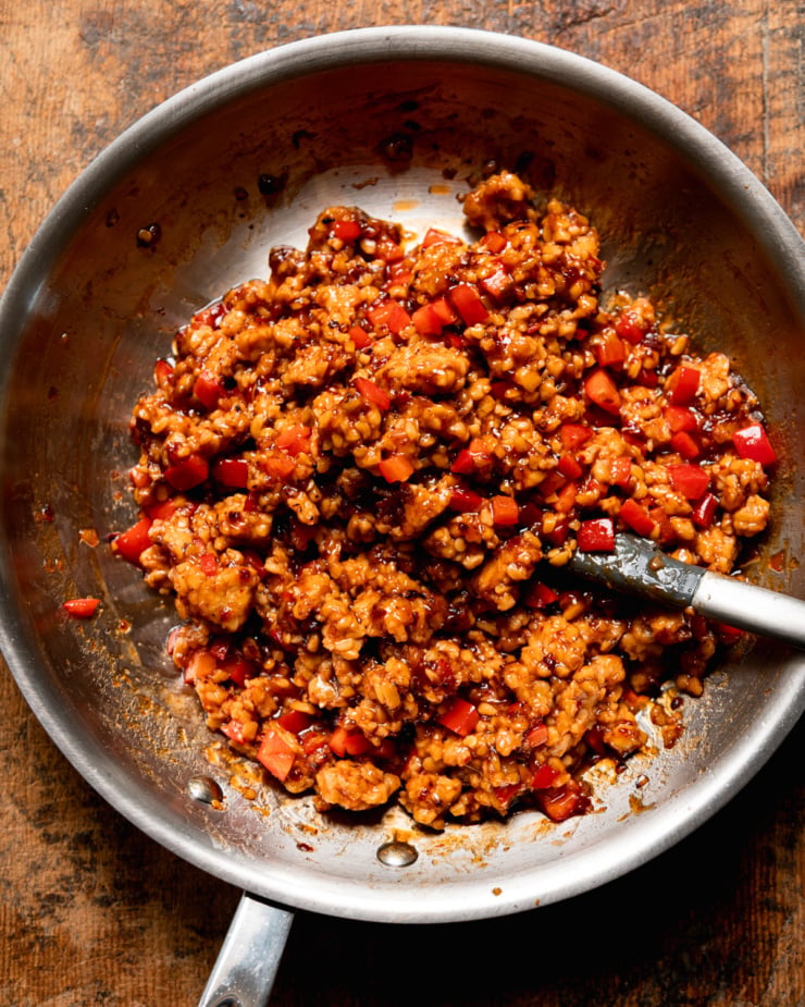 Overhead shot of a stainless steel skillet containing crumbled tempeh cooked in a glossy dark sauce with diced red bell pepper, a silicone spatula resting in the pan. The filling is deeply caramelized and saucy.