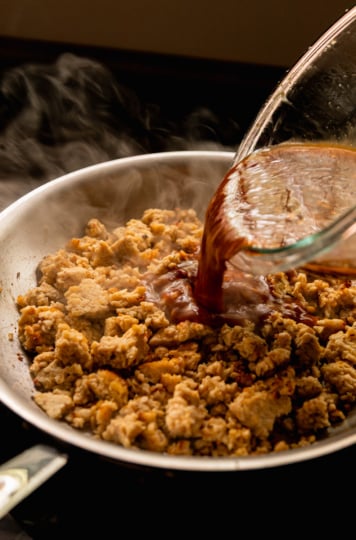 Action shot of a dark, glossy sauce being poured from a glass bowl into a hot stainless steel skillet of crumbled tempeh, with wisps of steam rising dramatically in the background.