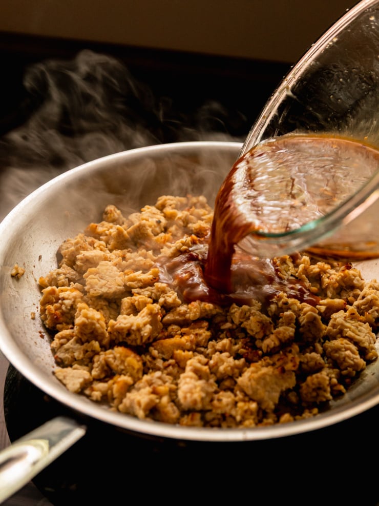 Action shot of a dark, glossy sauce being poured from a glass bowl into a hot stainless steel skillet of crumbled tempeh, with wisps of steam rising dramatically in the background.