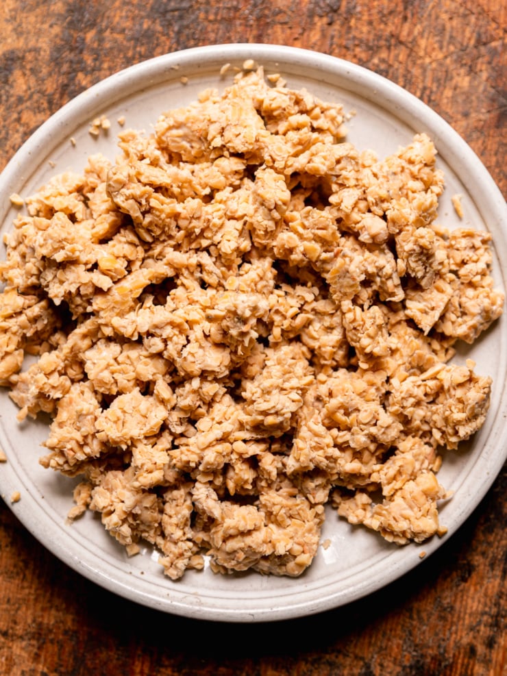 Overhead shot of crumbled, steamed tempeh on a round cream ceramic plate against a warm wood surface. The texture is chunky and irregular, pale golden in color.