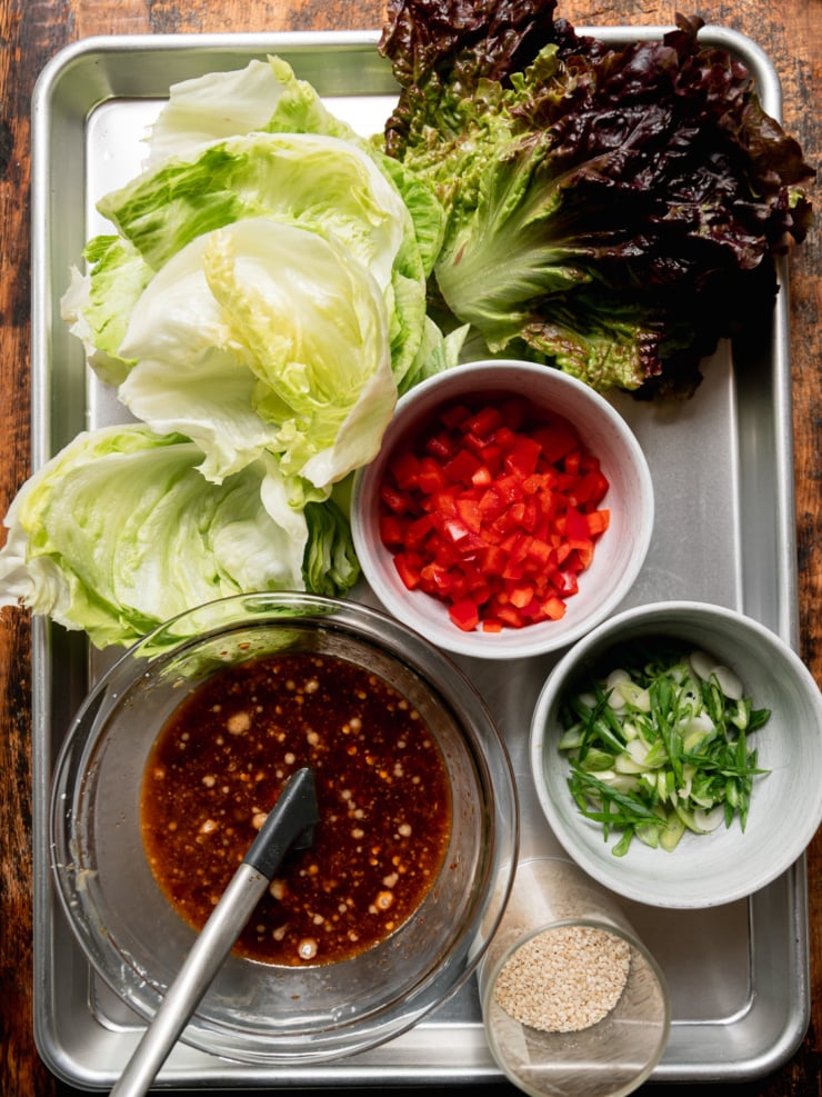 Overhead flat lay on a metal sheet pan showing prepped ingredients: iceberg lettuce cups, a head of dark red leaf lettuce, a small bowl of diced red bell pepper, a glass bowl of dark sauce with a spatula, a small bowl of sliced green onions, and a tiny dish of white sesame seeds.