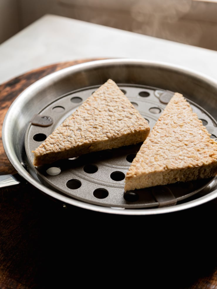 Side-angle shot of two triangular tempeh pieces on a metal steamer insert inside a pot, lit dramatically against a dark background. The tempeh's characteristic bean-and-grain texture is clearly visible.
