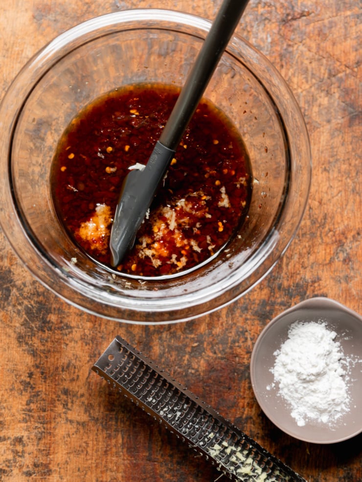 Overhead shot of a glass mixing bowl filled with dark glossy sauce — a blend of soy sauce, sesame oil, maple syrup, sambal oelek, rice vinegar, ginger, and garlic — being stirred with a silicone spatula. A microplane grater and a small dish of cornstarch sit alongside on a worn wood surface.