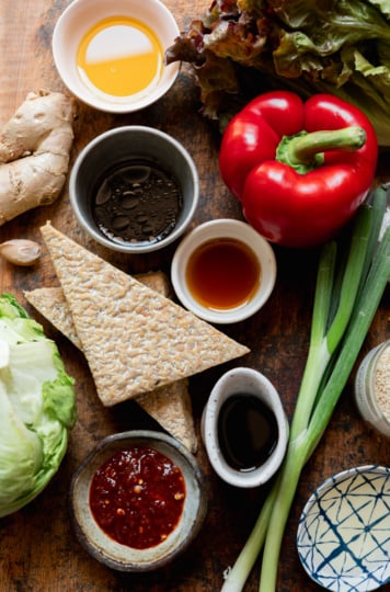 Overhead flat lay of all recipe ingredients on a warm wood surface: two triangles of tempeh, a head of iceberg lettuce, dark red leaf lettuce, a whole red bell pepper, fresh ginger root, a garlic clove, green onions, a bowl of sambal oelek, a small dish of soy sauce, a bowl of soy sauce), a bowl of sesame oil, a bowl of rice vinegar, a jar of sesame seeds, and a small dish of cornstarch.