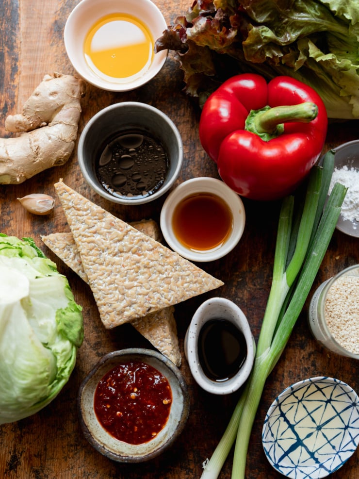 Overhead flat lay of all recipe ingredients on a warm wood surface: two triangles of tempeh, a head of iceberg lettuce, dark red leaf lettuce, a whole red bell pepper, fresh ginger root, a garlic clove, green onions, a bowl of sambal oelek, a small dish of soy sauce, a bowl of soy sauce), a bowl of sesame oil, a bowl of rice vinegar, a jar of sesame seeds, and a small dish of cornstarch.