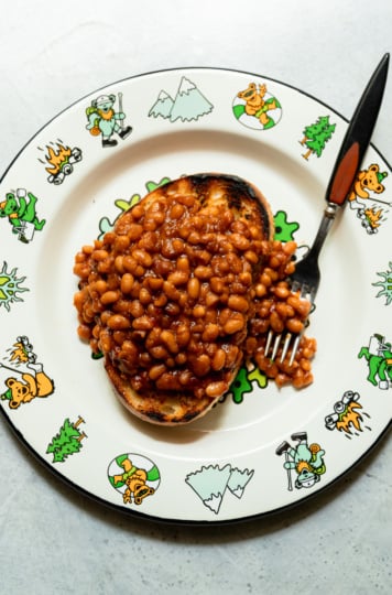 Overhead shot of saucy vegan maple baked beans piled on a thick slice of charred sourdough toast, served on a Grateful Dead-themed plate with a fork resting alongside.
