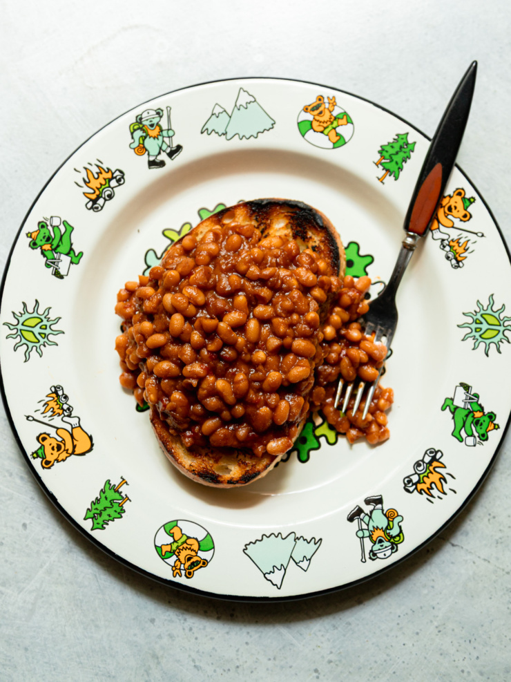 Overhead shot of saucy vegan maple baked beans piled on a thick slice of charred sourdough toast, served on a Grateful Dead-themed plate with a fork resting alongside.