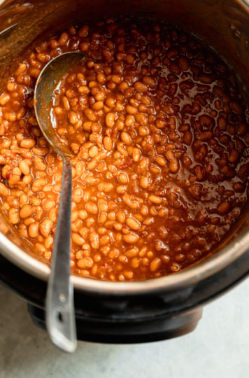 Overhead view of glossy, deep amber baked beans in the Instant Pot insert, partially stirred with a spoon resting in the pot.