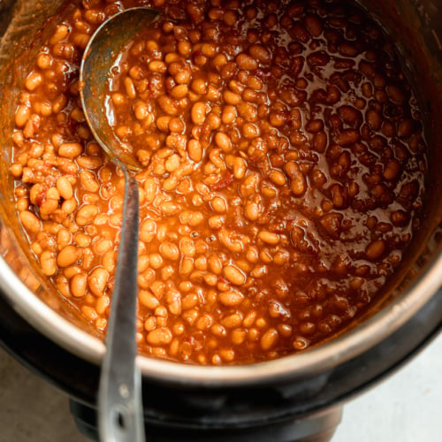 Overhead view of glossy, deep amber baked beans in the Instant Pot insert, partially stirred with a spoon resting in the pot.