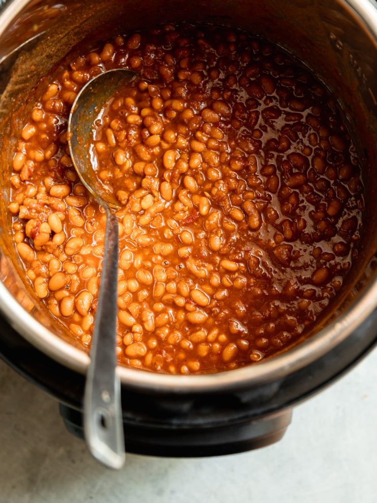 Overhead view of glossy, deep amber baked beans in the Instant Pot insert, partially stirred with a spoon resting in the pot.