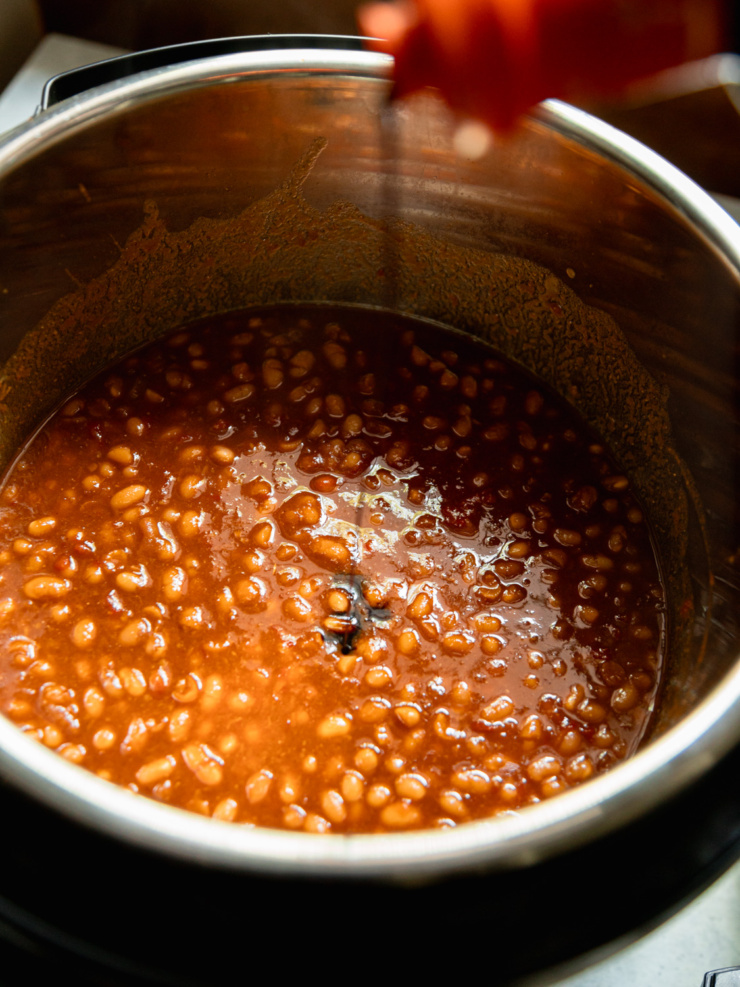 Overhead close-up of cooked white beans in their dark, glossy sauce inside the Instant Pot, with some balsamic vinegar being poured in.