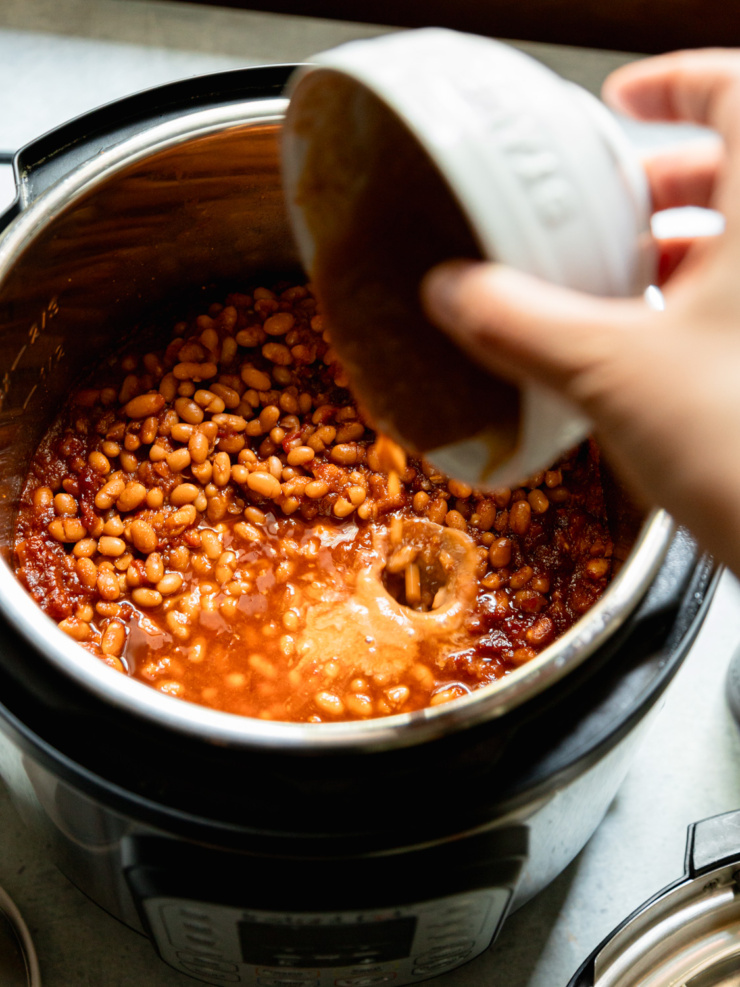 A hand tips a bowl of miso-enriched bean liquid into the Instant Pot, where they meet the rich tomato-maple sauce already inside.