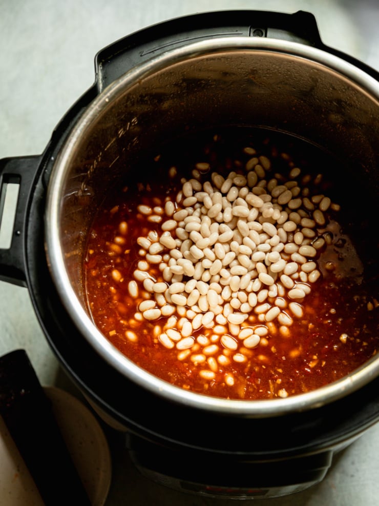 Overhead shot of pale, soaked white beans poured into the base of the bean sauce in the Instant Pot, not yet stirred together.