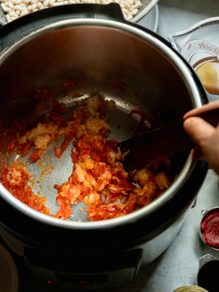 Overhead close-up of grated onion and red bell pepper softening in the bottom of the Instant Pot, with a wooden spatula stirring them.