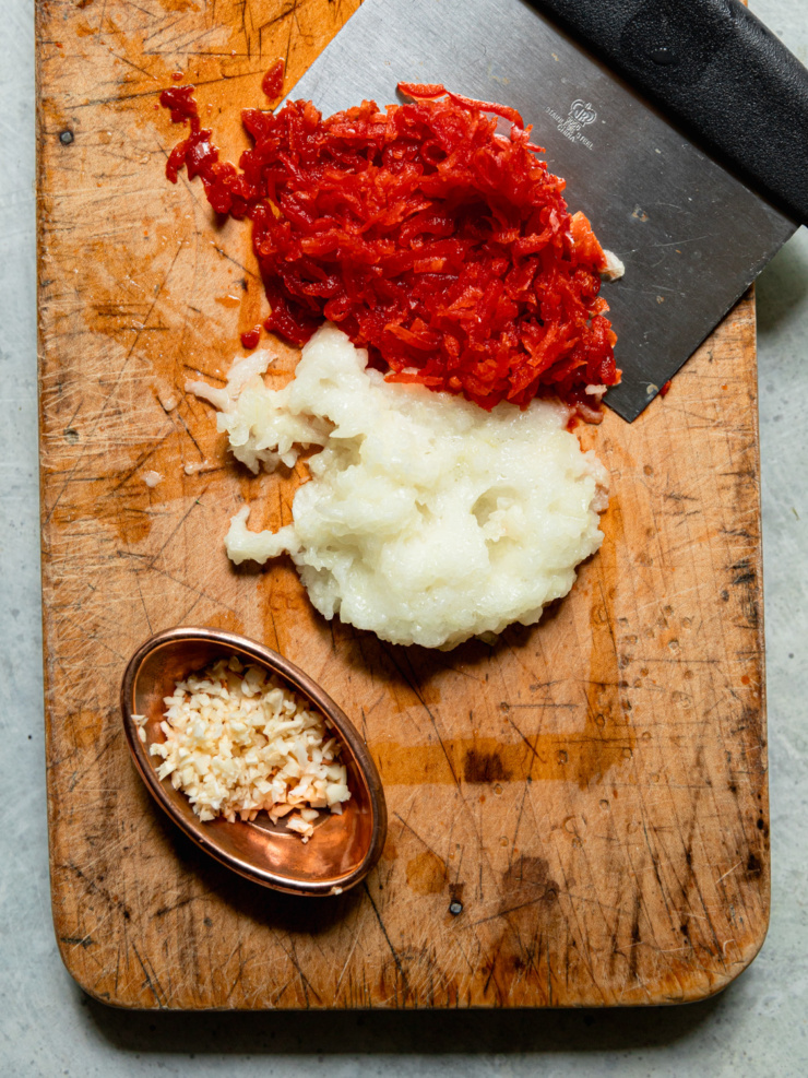 Overhead shot of grated red bell pepper, grated onion, and minced garlic arranged in separate piles on a well-worn wooden cutting board.