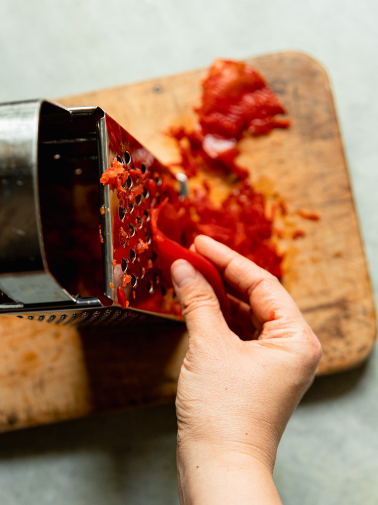 Close-up action shot of a hand grating red bell pepper on a box grater, with grated pepper piling up on the cutting board below.